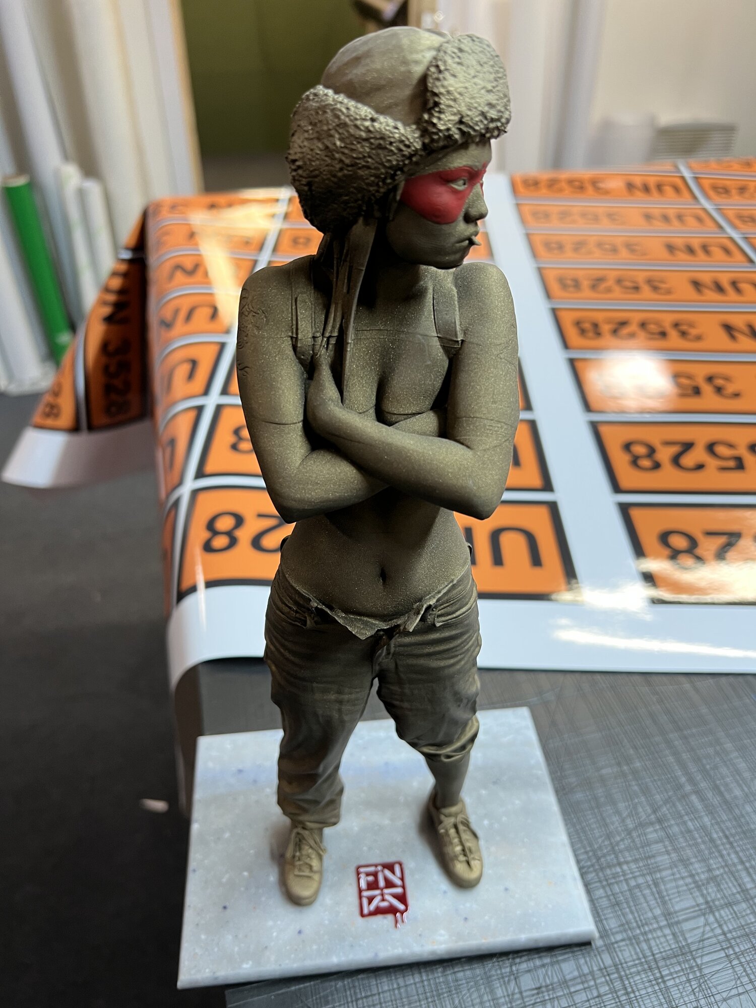 Sand-cast bronze finish with hand-painted red mask — packed and ready alongside hazard-marked transit boards.